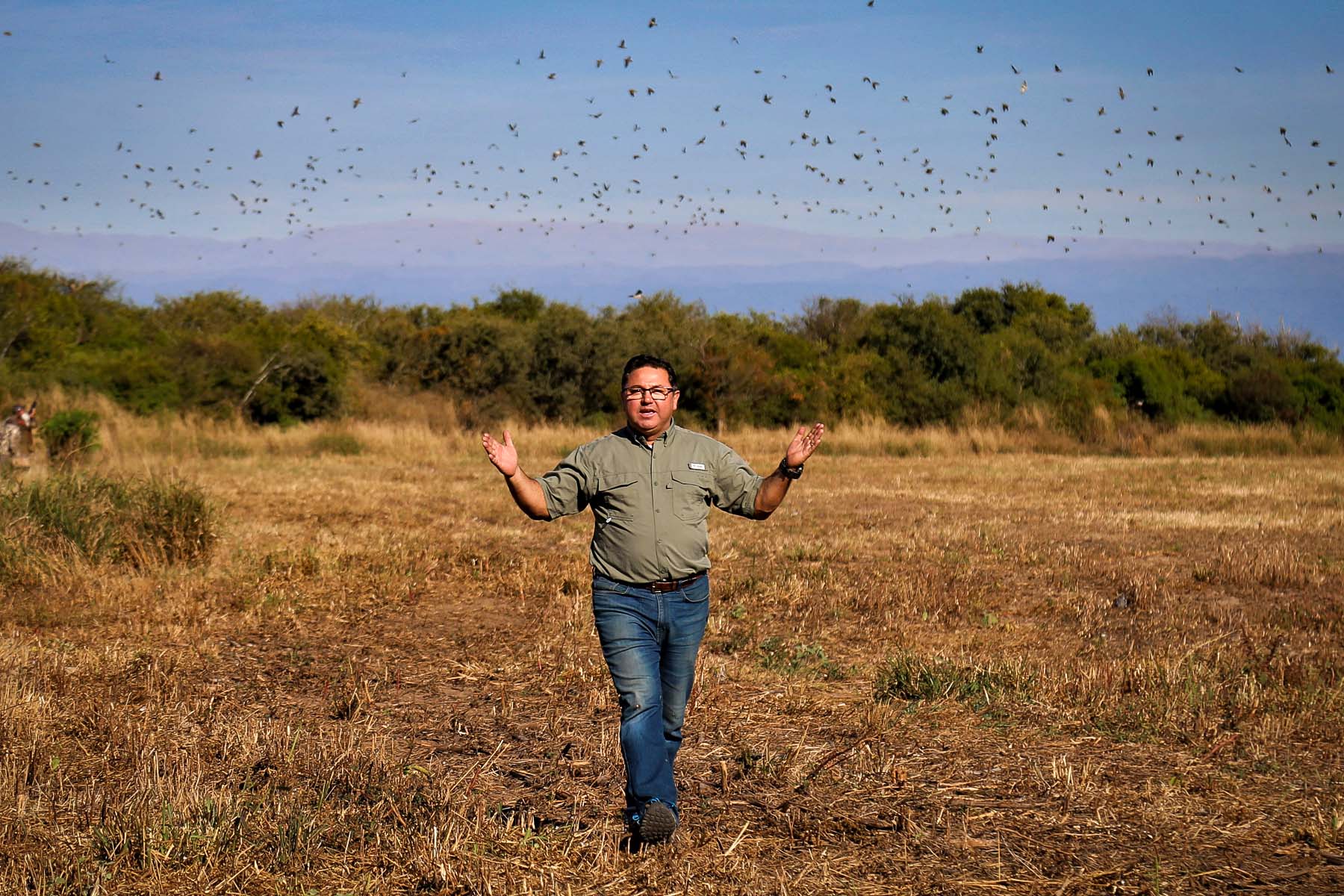 Nacho, owner of C&C Outfitters, walking with arms wide open in the field, surrounded by a vast landscape and hundreds of doves flying in the sky at Alijilan Lodge, Argentina